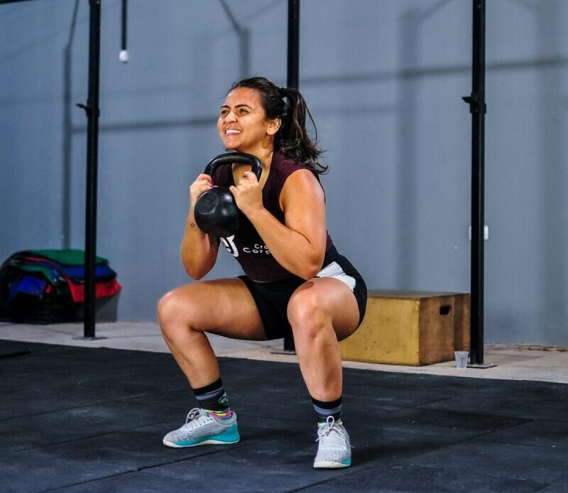 Smiling woman doing kettlebell squats indoors with a Brazilian flag in the background.